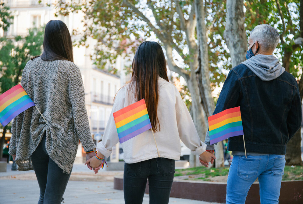 Drie mensen hand in hand op straat, op de rug gezien met LGBTIQ+-vlaggetje in hun achterzak.