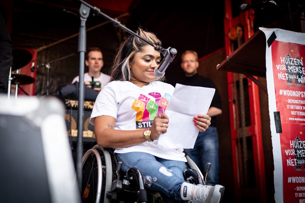 Een foto van Jeanette Chedda, enigszins van onderen genomen terwijl zij in haar rolstoel voor een microfoon zit. Ze draagt blauwe jeans en een wit T-shirt met kleurrijke opgestoken vuisten. Ze leest voor van een A4-papier.