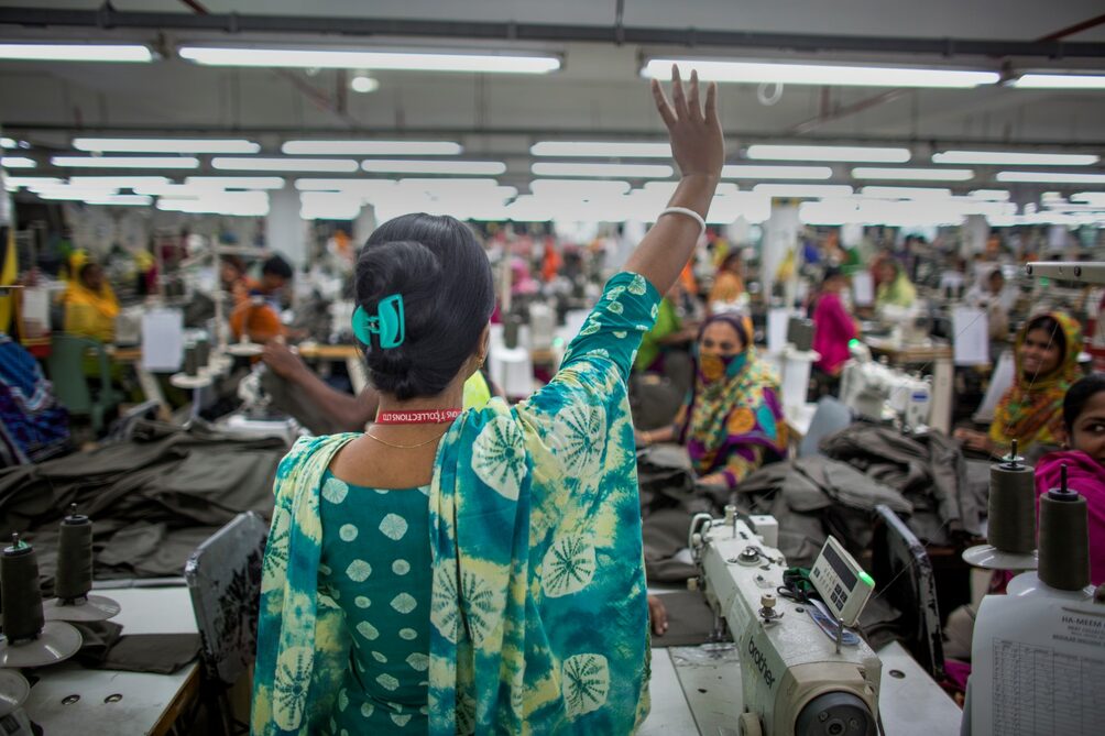 Een foto van een vrouw in een werkplaats voor kleding. Ze heeft een kleurrijke groene en gele jurk aan. Ze steekt haar rechterhand op. Op de achtergrond zitten en staan allemaal arbeiders te werken in de drukke werkplaats.