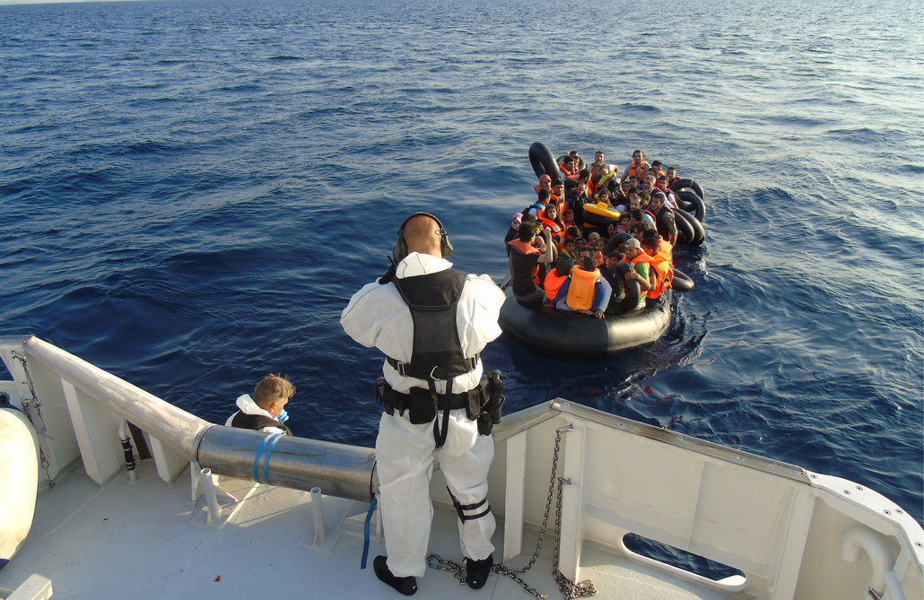 Twee personen in witte pakken staan op een hoge witte boot. Ze staan met hun rug naar de camera en ze kijken naar een klein zwart rubberbootje vol met mensen. Deze mensen hebben allemaal reddingsvesten aan en zwembanden vast.